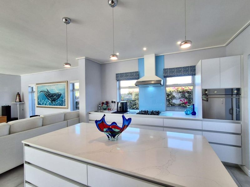 Modern open-plan kitchen featuring a large central island with quartz countertops, handleless white cabinetry, built-in oven, and gas hob with a blue glass splashback.