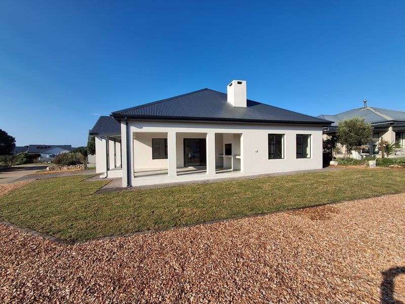 Rear view of a modern house featuring a covered patio with a built-in braai, corrugated metal roof, and well-maintained lawn and gravel landscaping.