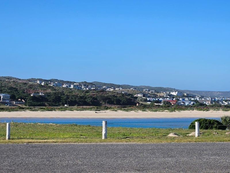 Scenic coastal view overlooking a sandy beach, estuary, and hillside residential area from a roadside vantage point.