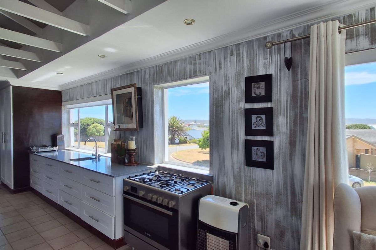 A bright kitchen featuring a gas cooktop, stainless steel countertops, and large windows offering scenic views, complemented by exposed ceiling beams and tiled flooring.
