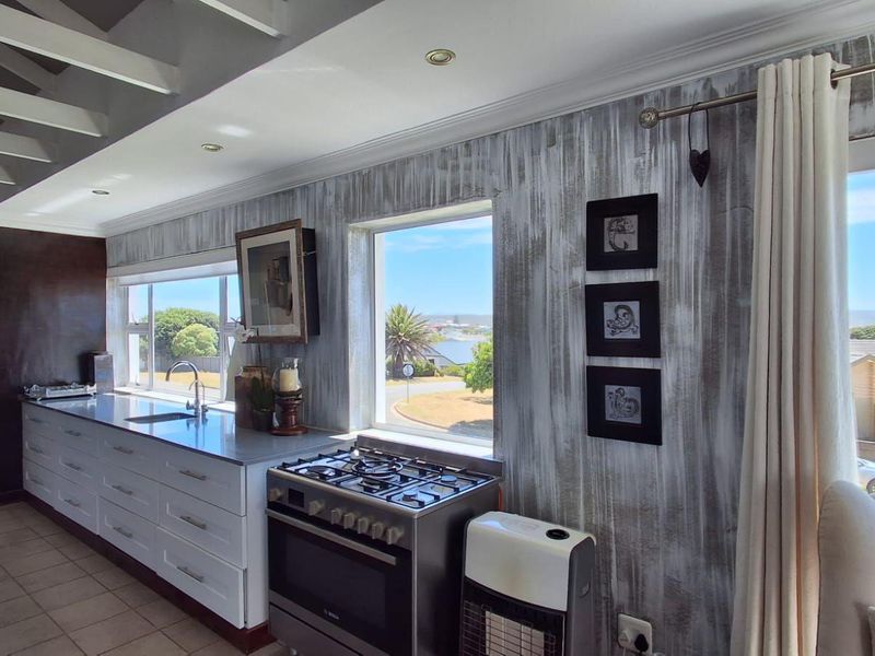 A bright kitchen featuring a gas cooktop, stainless steel countertops, and large windows offering scenic views, complemented by exposed ceiling beams and tiled flooring.