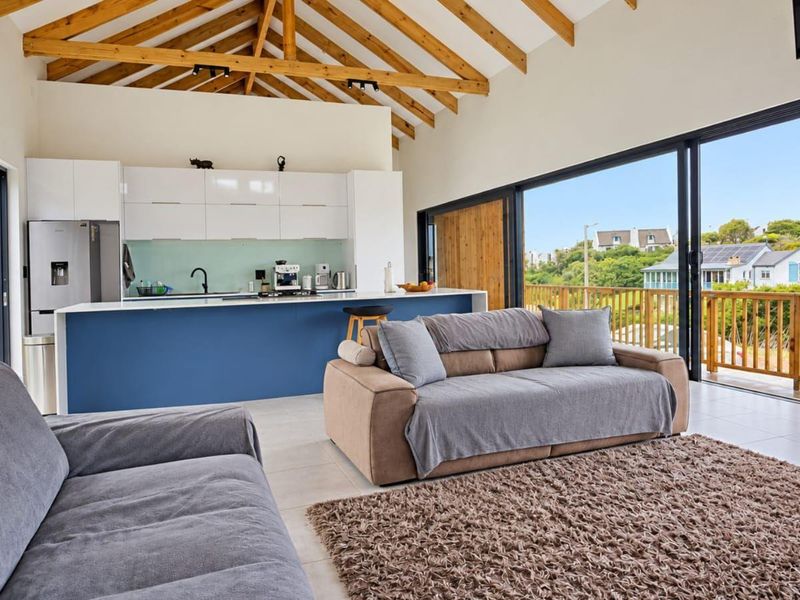 Modern open-plan living area featuring a sleek kitchen with island, exposed timber trusses, and large sliding doors opening to a balcony with scenic views.