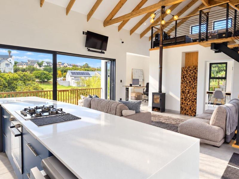 Spacious open-plan living area featuring a modern kitchen island, wood-burning fireplace, exposed timber rafters, and a mezzanine level with access to a balcony.