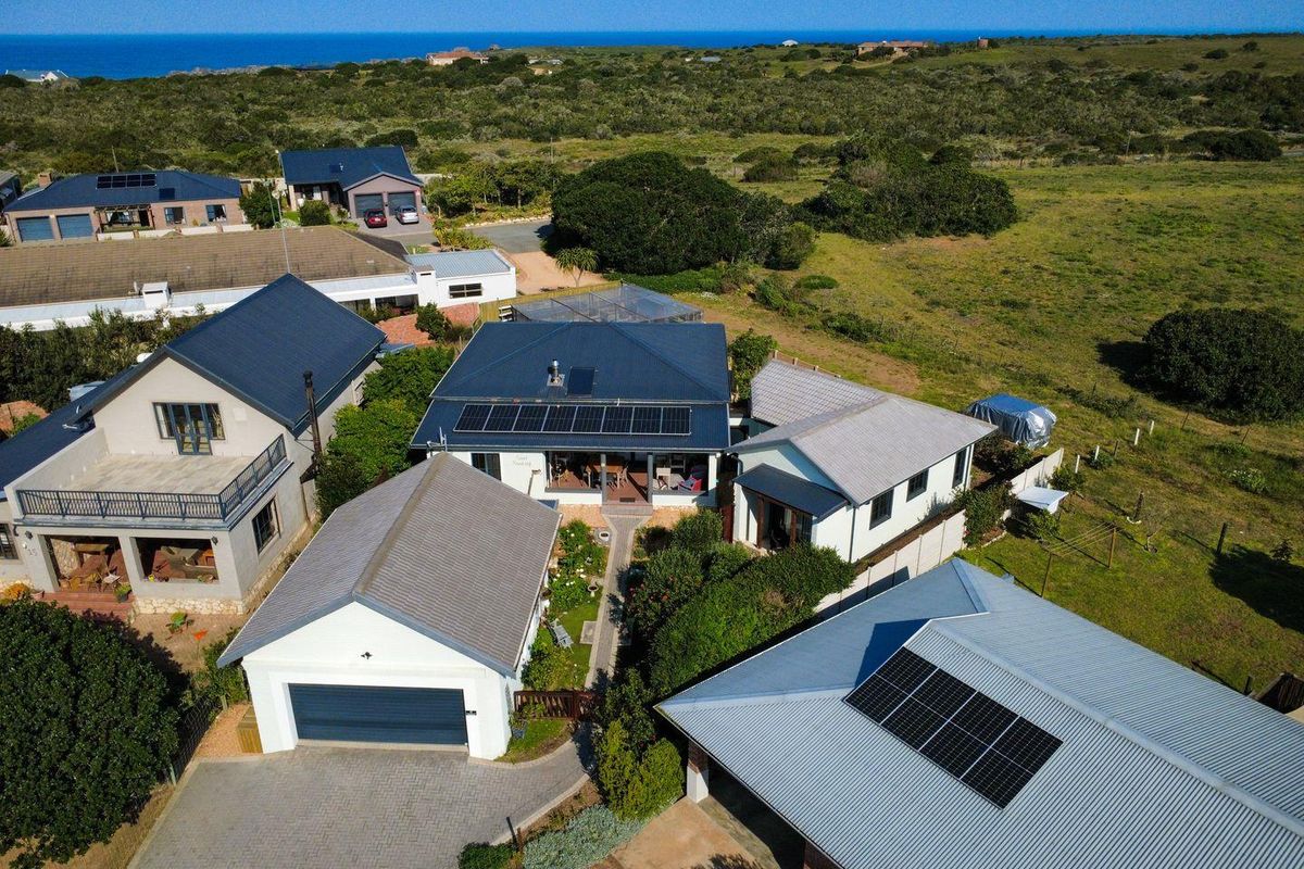 Aerial view of a residential property featuring multiple buildings with metal roofs, solar panels, a paved driveway, and surrounding coastal landscape.