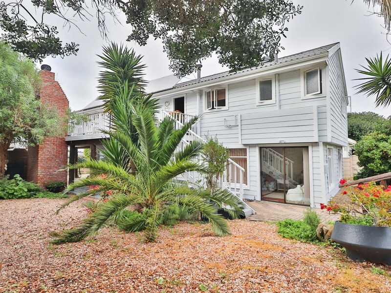Rear view of a two-story weatherboard house featuring a raised timber deck, external staircase, brick chimney, and a paved patio area set within a garden.