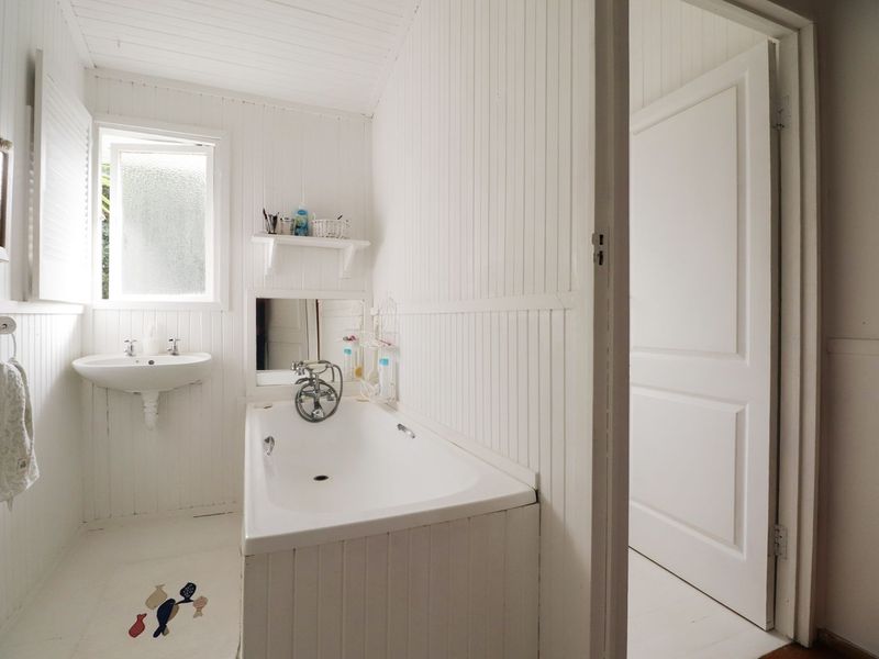 A compact bathroom featuring white beadboard wall paneling, a built-in bathtub, and a wall-mounted basin with natural light from an obscured glass window.