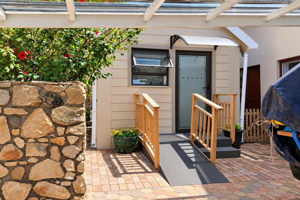 A well-maintained rear exterior area featuring a stone retaining wall, paved driveway, and a covered entrance with an accessibility ramp and timber handrails.