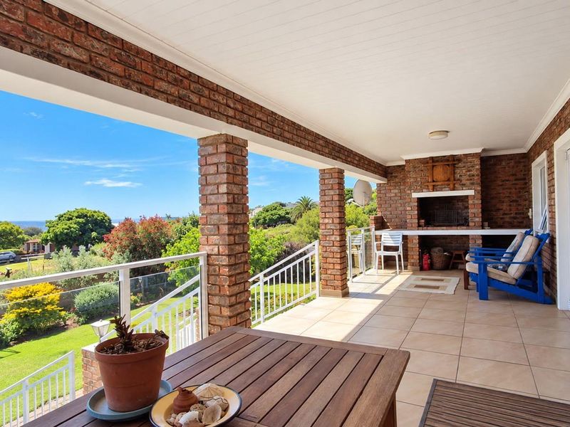 Spacious covered patio featuring a built-in brick braai, tiled flooring, and a glass balustrade with scenic garden and coastal views.