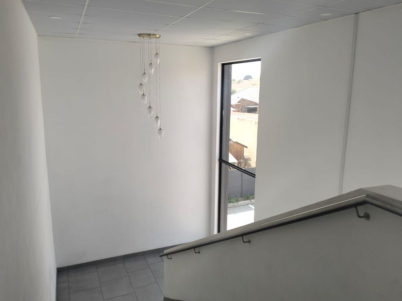 A bright, minimalist office hallway featuring a staircase with a metal handrail, tiled flooring, and a large window providing natural light.