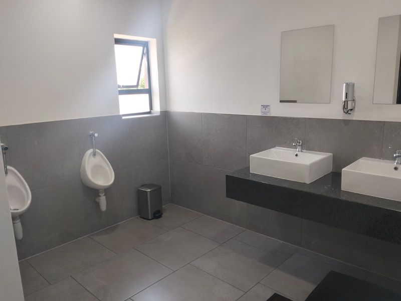 Modern office bathroom featuring dual countertop basins on a floating granite vanity, wall-mounted urinals, and neutral grey floor-to-wall tiling.