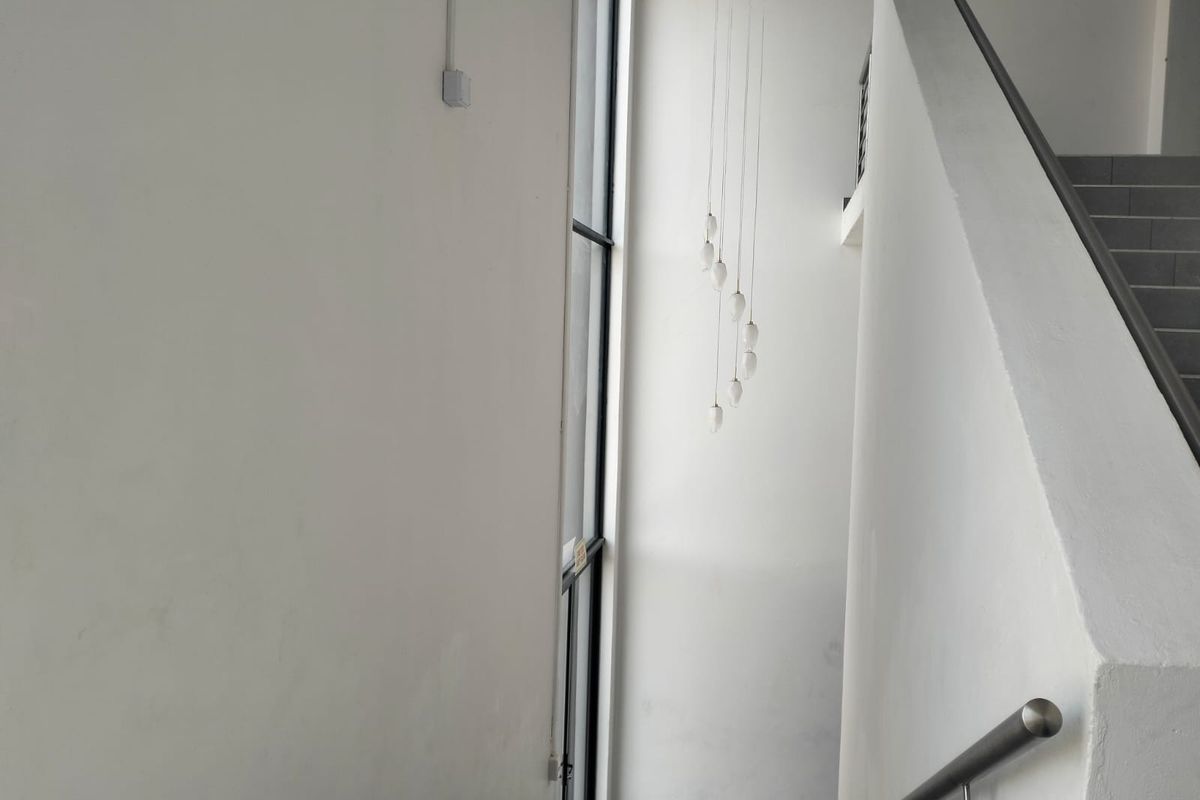 A bright, minimalist office hallway featuring a staircase with a metal handrail, large floor-to-ceiling windows, and modern pendant lighting.