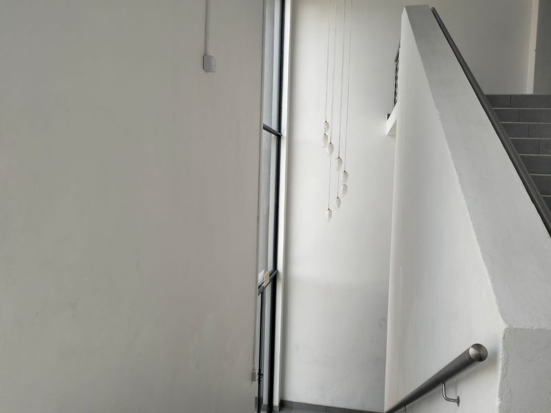 A bright, minimalist office hallway featuring a staircase with a metal handrail, large floor-to-ceiling windows, and modern pendant lighting.