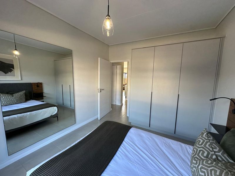 A modern bedroom featuring neutral tones, built-in cupboards, a large wall mirror, and tiled flooring, offering a clean and contemporary living space.
