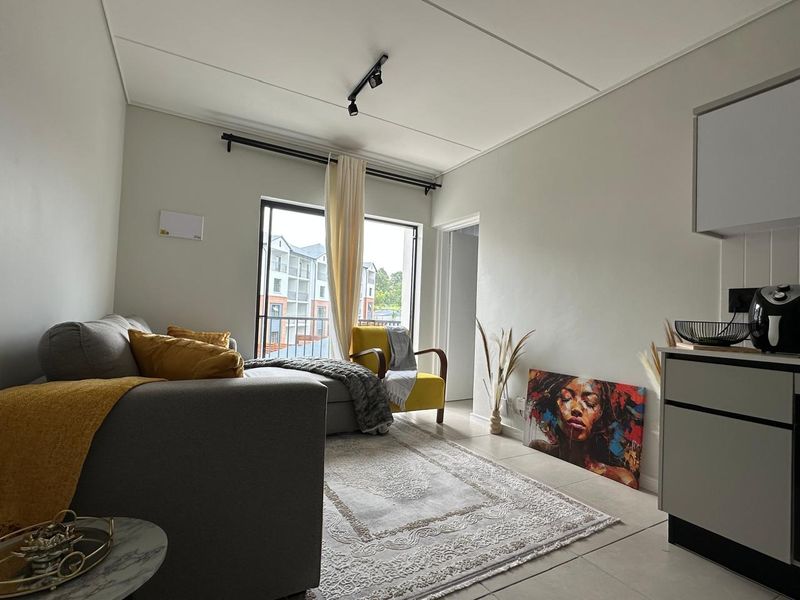 An open-plan living area featuring tiled floors, modern track lighting, and a sliding door leading to a balcony, integrated with a compact kitchen space.