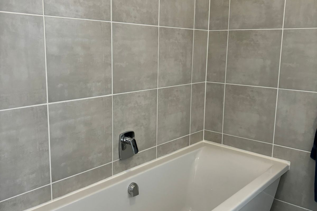 A clean, modern bathroom featuring a built-in bathtub with contemporary chrome fixtures and neutral grey wall tiling.