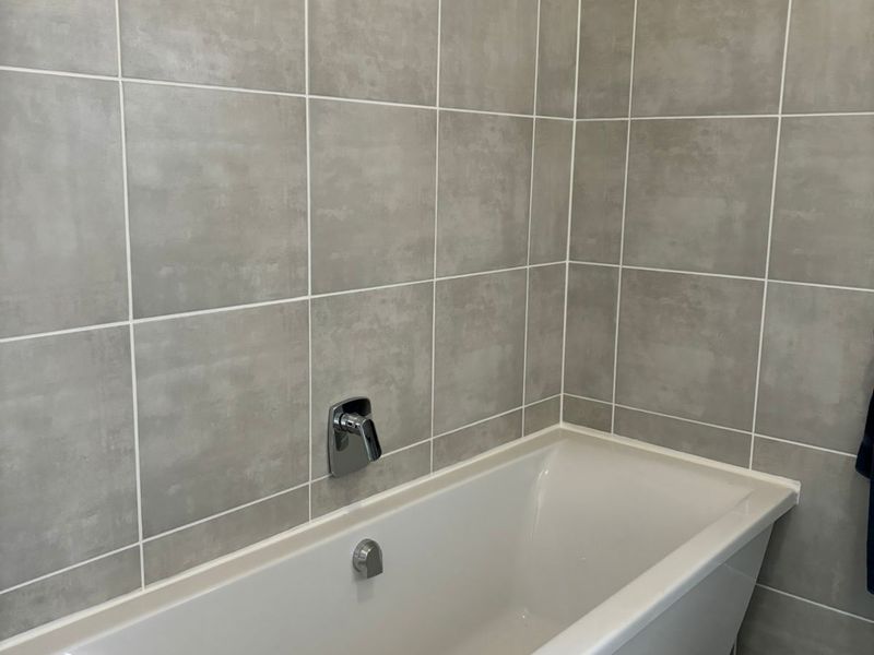A clean, modern bathroom featuring a built-in bathtub with contemporary chrome fixtures and neutral grey wall tiling.
