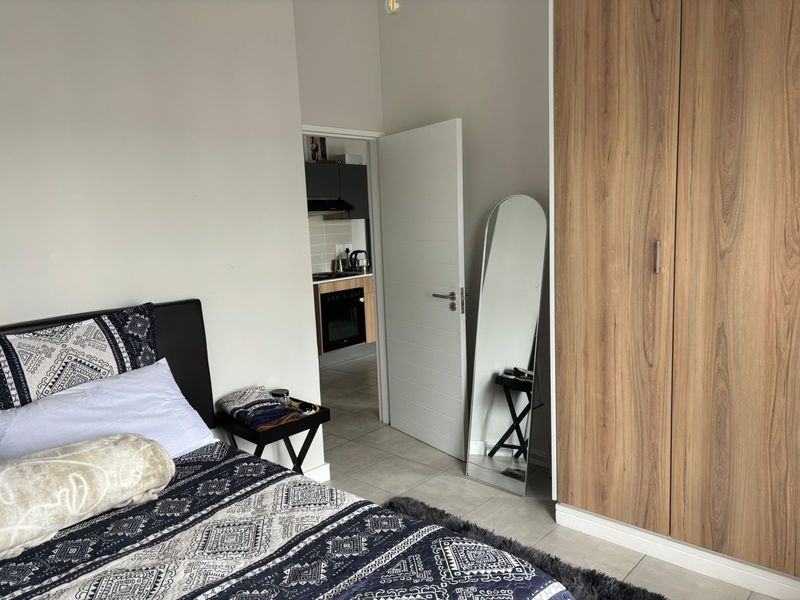 A well-maintained bedroom featuring modern wood-finish built-in wardrobes, neutral wall tones, and tiled flooring, with a view into the adjacent kitchen area.