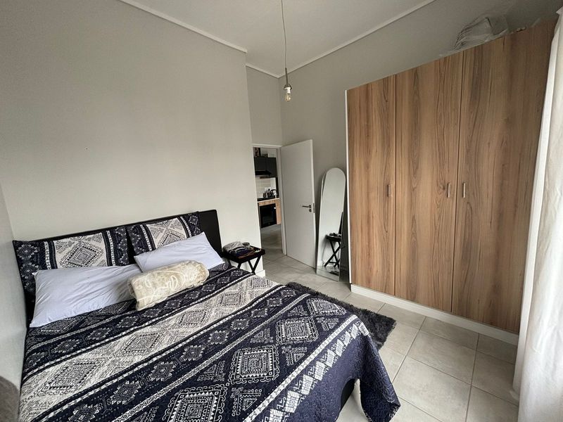 A well-maintained bedroom featuring neutral walls, tiled flooring, and a large built-in wooden wardrobe, with a view into the adjacent kitchen area.