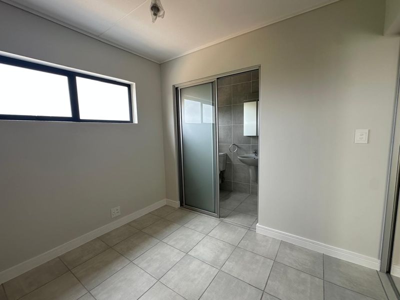 A clean, neutral bedroom featuring tiled flooring, a horizontal window, and an en-suite bathroom accessed via a sliding door.