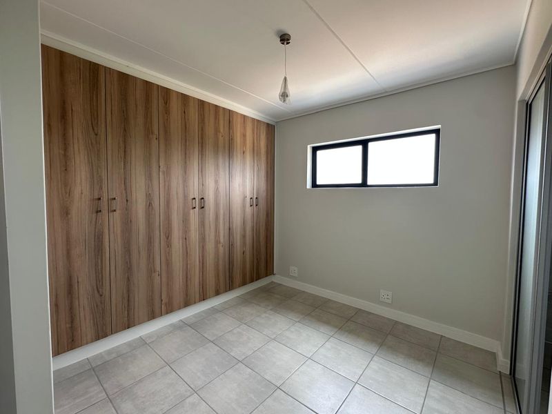 A clean, modern bedroom featuring neutral tiled flooring, light grey walls, and extensive built-in wood-grain cabinetry for ample storage.