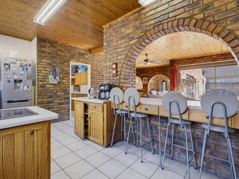 A rustic kitchen featuring exposed brick walls, timber ceiling, tiled flooring, and a breakfast bar opening into the adjacent living area.