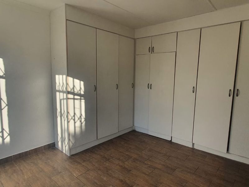 A bright, empty bedroom featuring extensive built-in cupboard storage, tiled flooring, and a security gate on the window.