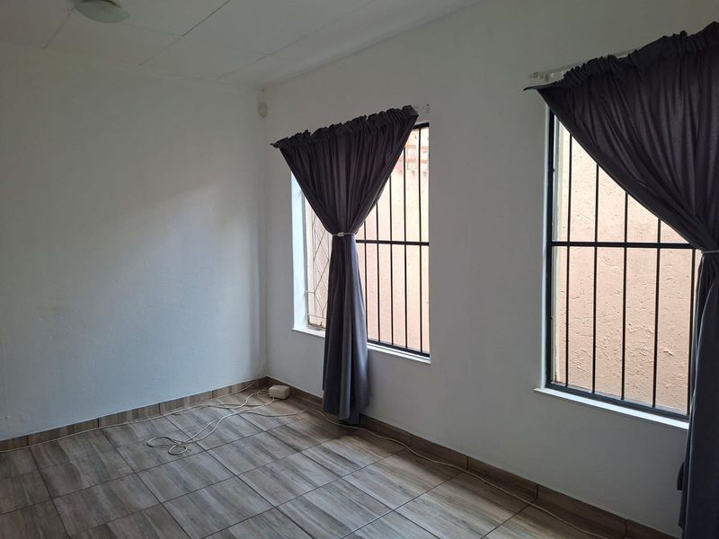 A bright, empty bedroom featuring wood-look tiled flooring and windows fitted with security bars.