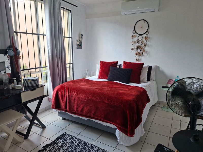 A functional bedroom featuring tiled flooring, a wall-mounted split-system air conditioner, and large windows fitted with security bars.
