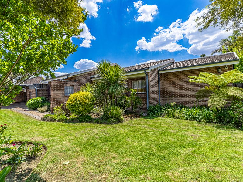 A well-maintained rear exterior view of the house featuring a classic brick facade, tiled roof, and a lush, landscaped garden with a paved walkway.