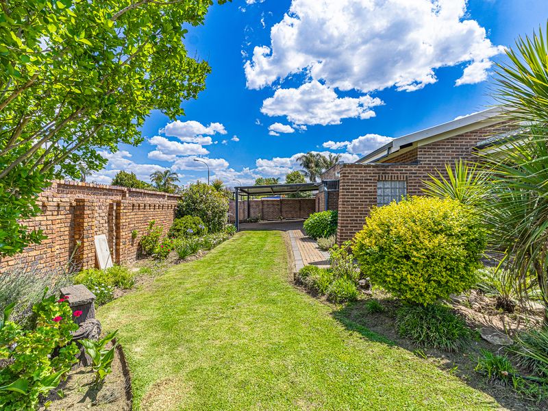 A well-maintained rear garden featuring a lush lawn, established landscaping, brick boundary walls, and a paved path leading to a rear carport.