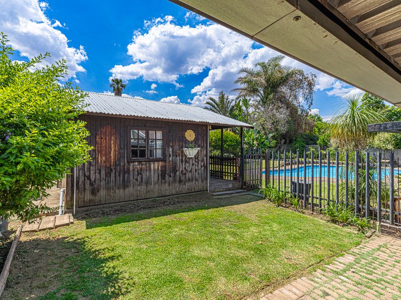 Rear garden view featuring a timber-clad outbuilding, a swimming pool enclosed by metal fencing, and a paved walkway under a covered patio area.