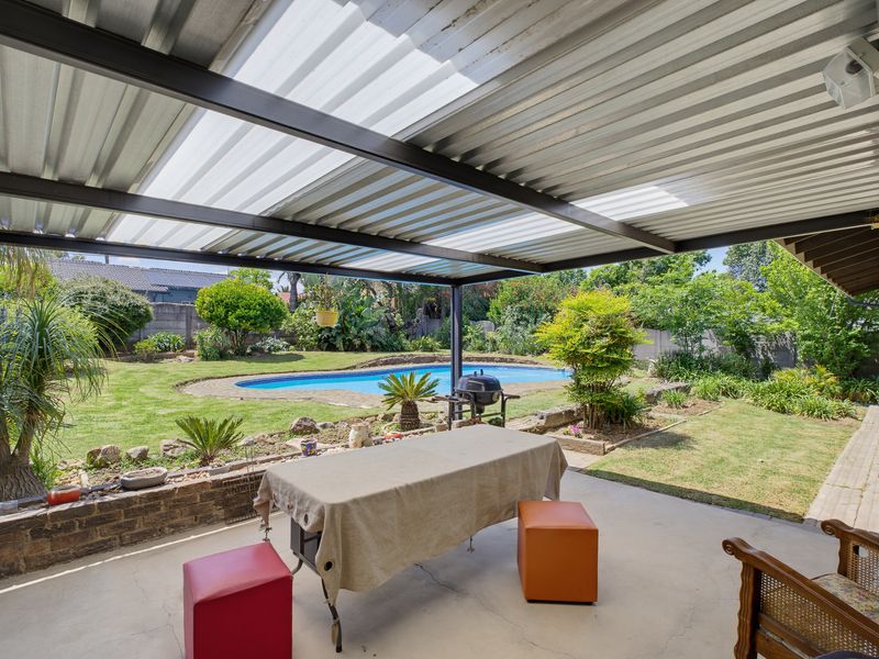 Covered outdoor patio area featuring a corrugated metal roof with translucent panels, overlooking a landscaped garden and swimming pool.