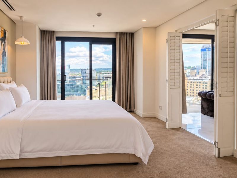 Spacious bedroom featuring neutral carpeting, recessed lighting, ducted air conditioning, and sliding glass doors leading to a private balcony with city views.