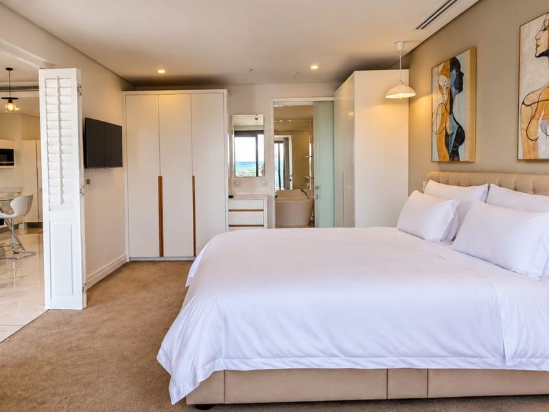 Spacious bedroom featuring built-in cupboards, neutral carpeting, recessed lighting, and direct access to an en-suite bathroom and adjacent kitchen area.