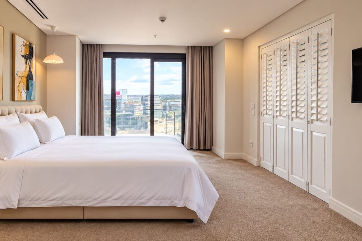 Spacious bedroom featuring built-in cupboards with shutter doors, ducted air conditioning, and a sliding glass door leading to a balcony with city views.