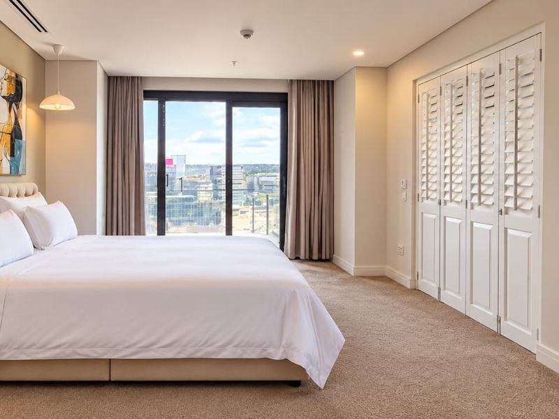 Spacious bedroom featuring built-in cupboards with shutter doors, ducted air conditioning, and a sliding glass door leading to a balcony with city views.