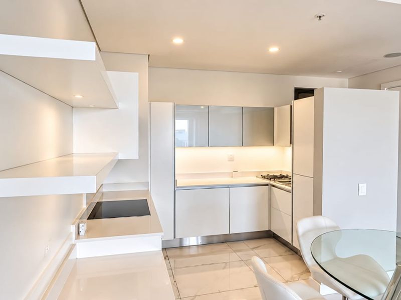 Modern open-plan kitchen featuring sleek white cabinetry, stone countertops, integrated cooktop, and floating shelving, complemented by recessed lighting and polished tiled floors.
