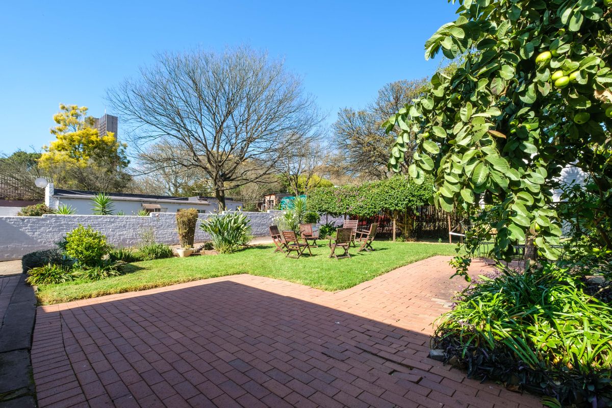 A spacious rear garden featuring a brick-paved patio area, a well-maintained lawn, and established perimeter walls with mature trees.