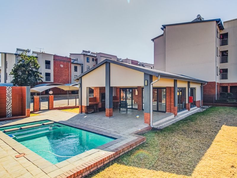 Communal pool area featuring a covered patio with built-in braai facilities, paved decking, and well-maintained brick and plaster architecture.