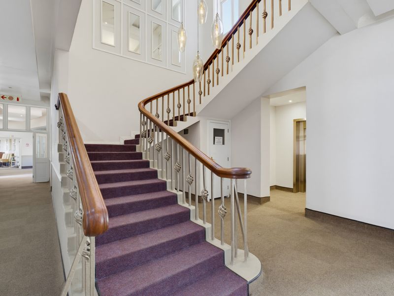 A grand entrance hallway featuring a sweeping staircase with a wooden handrail, carpeted flooring, and access to an elevator.