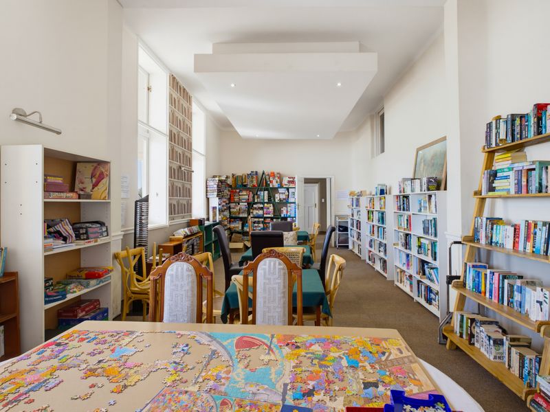 A spacious communal living area featuring high ceilings, recessed lighting, and extensive built-in shelving, currently utilized as a library and activity room.