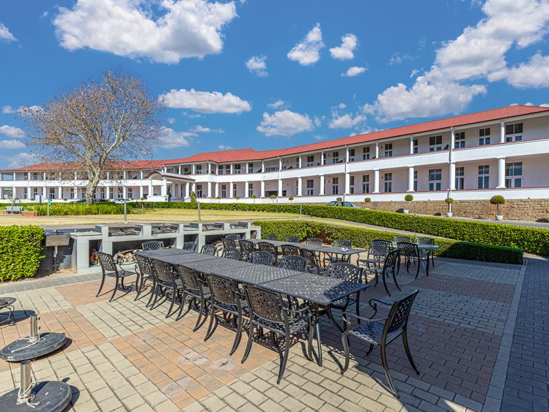 Spacious outdoor patio area featuring a built-in braai, paved flooring, and well-maintained garden hedges, set against a large apartment building facade.