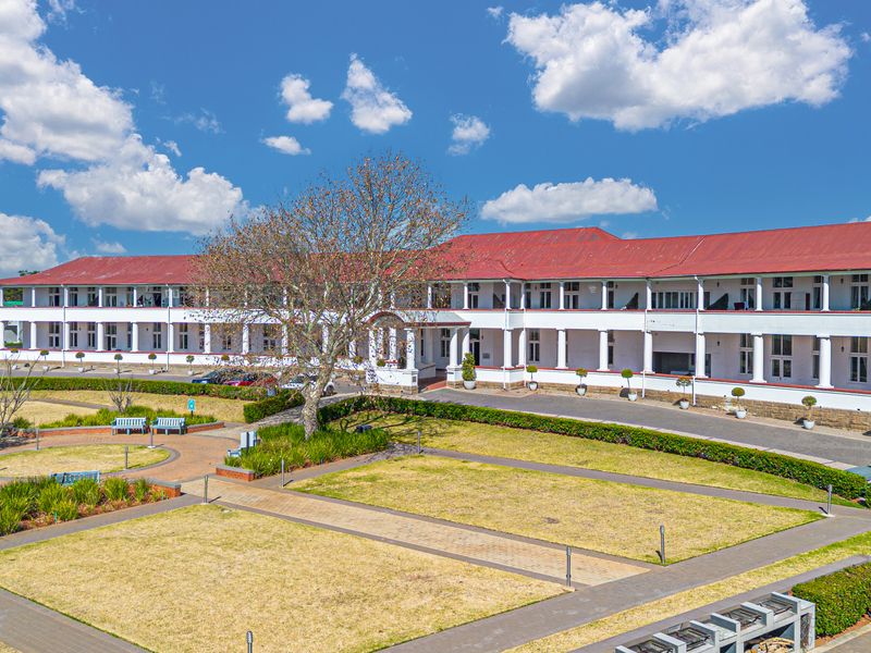 A wide-angle view of the apartment complex exterior, featuring a long covered veranda, metal roofing, and well-maintained landscaped grounds with paved walkways.