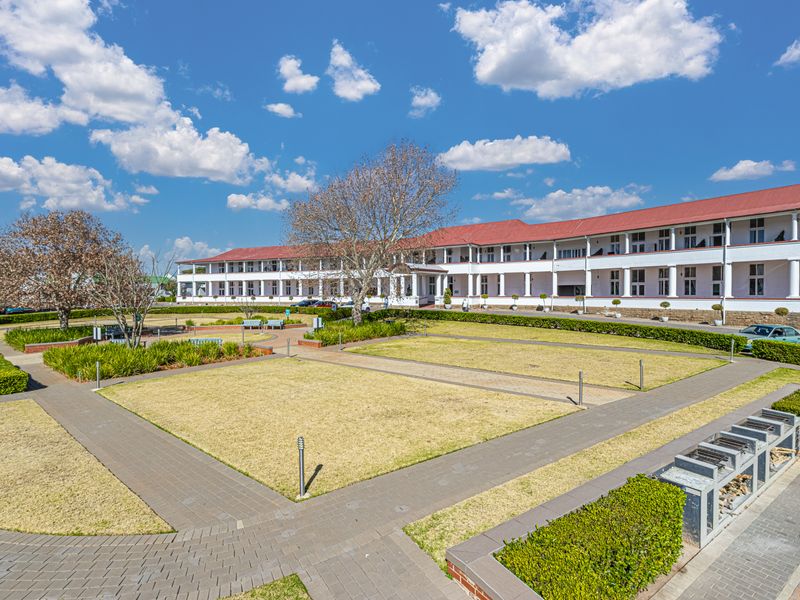 A well-maintained apartment complex exterior featuring a landscaped garden, paved walkways, and a covered veranda with built-in braai facilities.