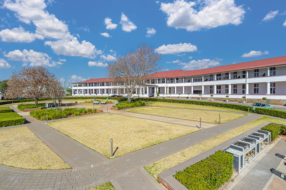 A well-maintained apartment complex exterior featuring a landscaped garden, paved walkways, and a covered veranda with built-in braai facilities.