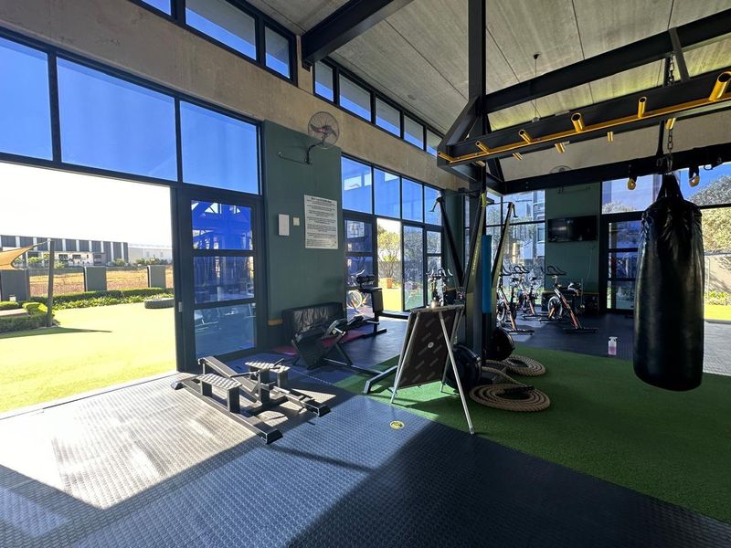 A modern, well-lit gym facility featuring high ceilings, exposed steel beams, large glass windows, and a combination of rubber and artificial turf flooring.