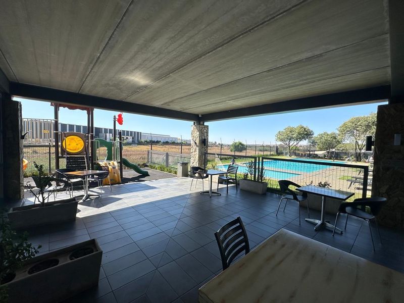 A spacious covered patio featuring tiled flooring and stone-clad pillars, overlooking a communal swimming pool and playground area.