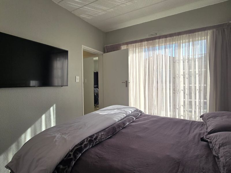 A well-lit bedroom featuring neutral wall tones, a wall-mounted television, and large windows with sheer curtains allowing for natural light.