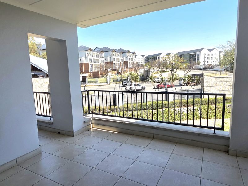 A spacious, covered balcony featuring tiled flooring and a metal balustrade, offering an open view of the surrounding residential complex.