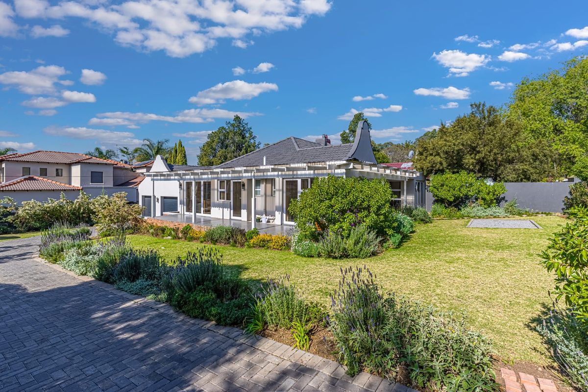 Rear view of the property featuring a well-maintained garden, paved driveway, and a covered patio area with sliding glass doors.